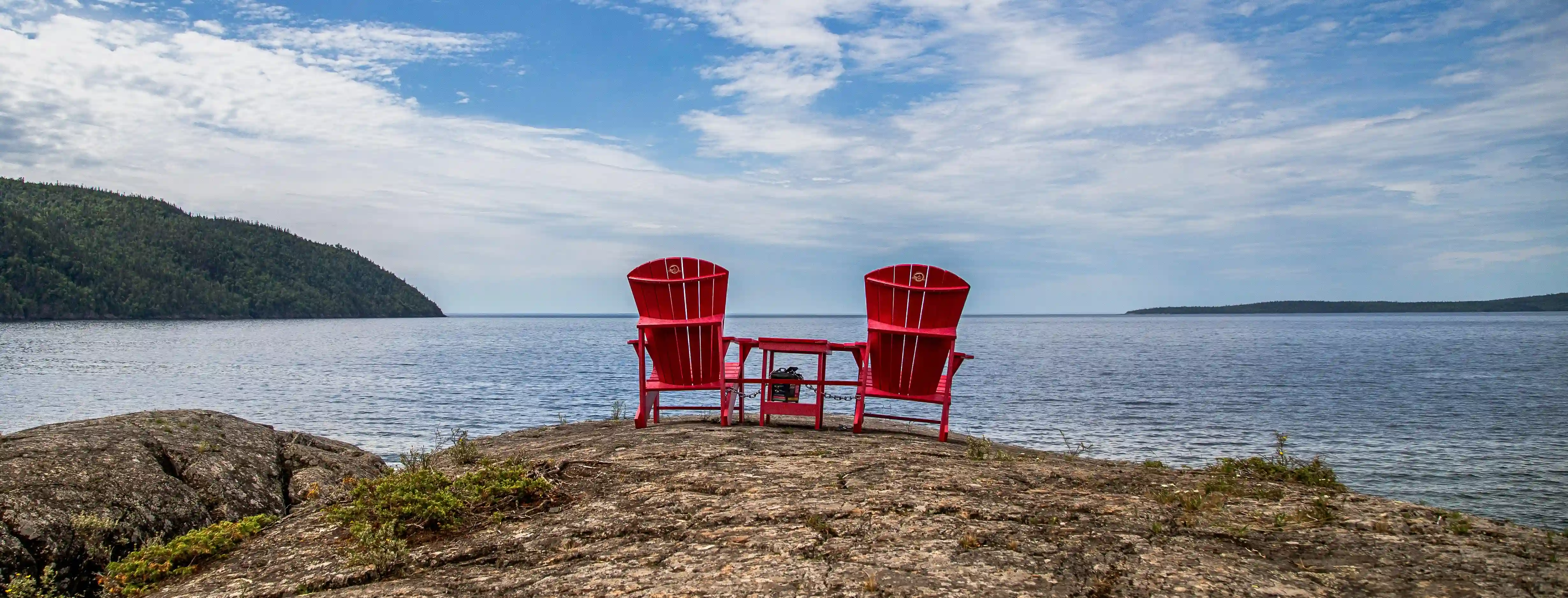 Two red chairs and table on the edge of the coast looking out to a blue sea and sky